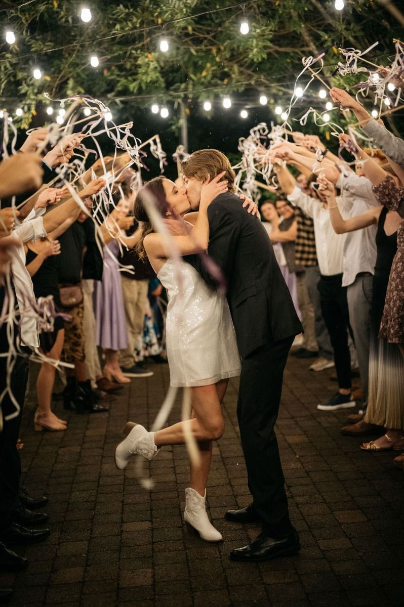 Couple kissing under string lights with guests holding white ribbons.