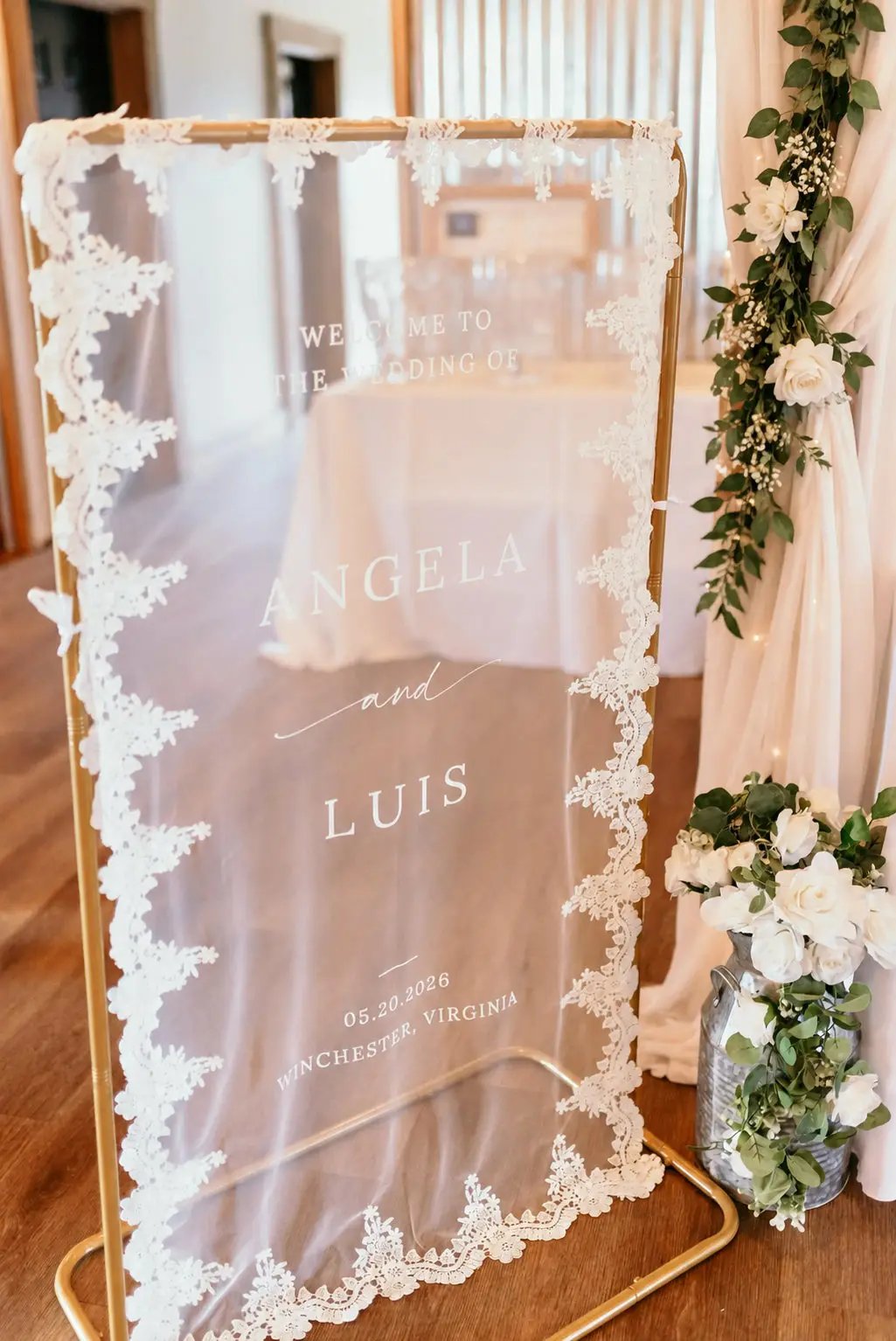 A wedding welcome sign with a gold metal frame and white lace trim, displayed on a wooden floor in a decorated event space with floral arrangements and draped fabric.
