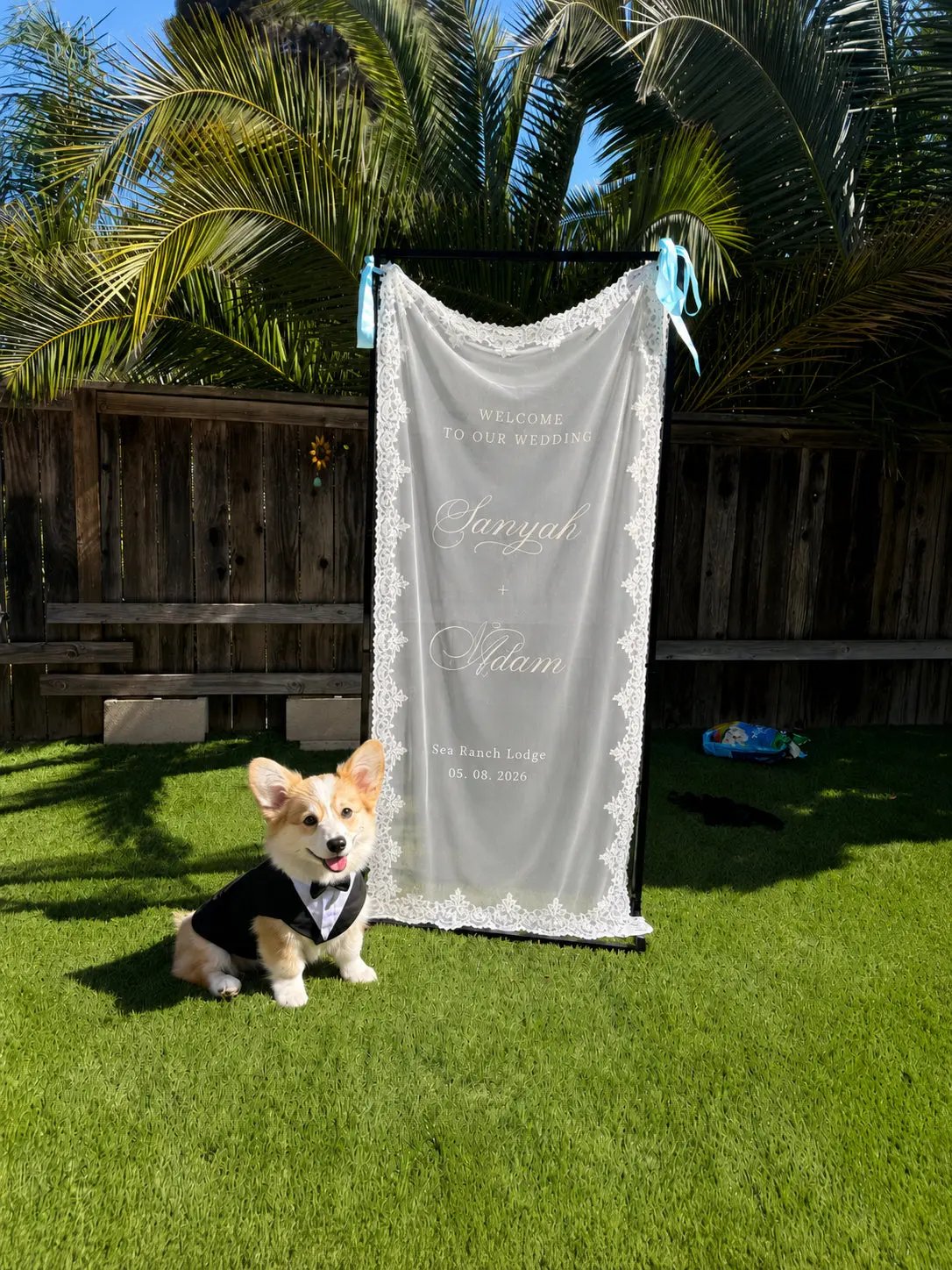 A small tan and white dog wearing a black bow tie sits on green grass in front of a custom white lace-edged wedding backdrop banner that reads 'Savvyul & Adam' with wedding details. The scene is outdoors on a wooden fence with palm trees in the background.